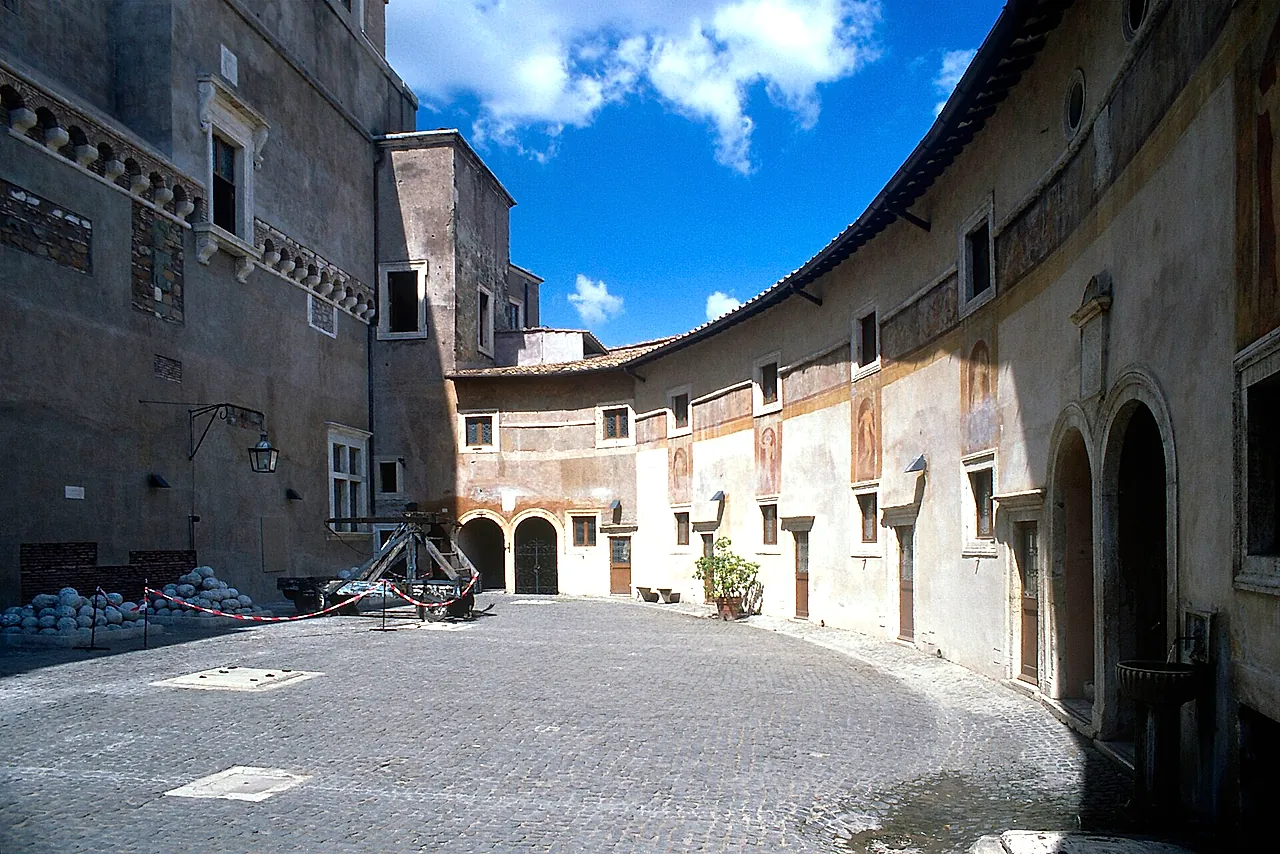 Der gebogene Hof Alexanders VI. in der Burg, mit verblassten Fresken an den Wänden, einem Haufen Steinkugeln und einem hölzernen Katapult, unter blauem Himmel mit einigen Wolken