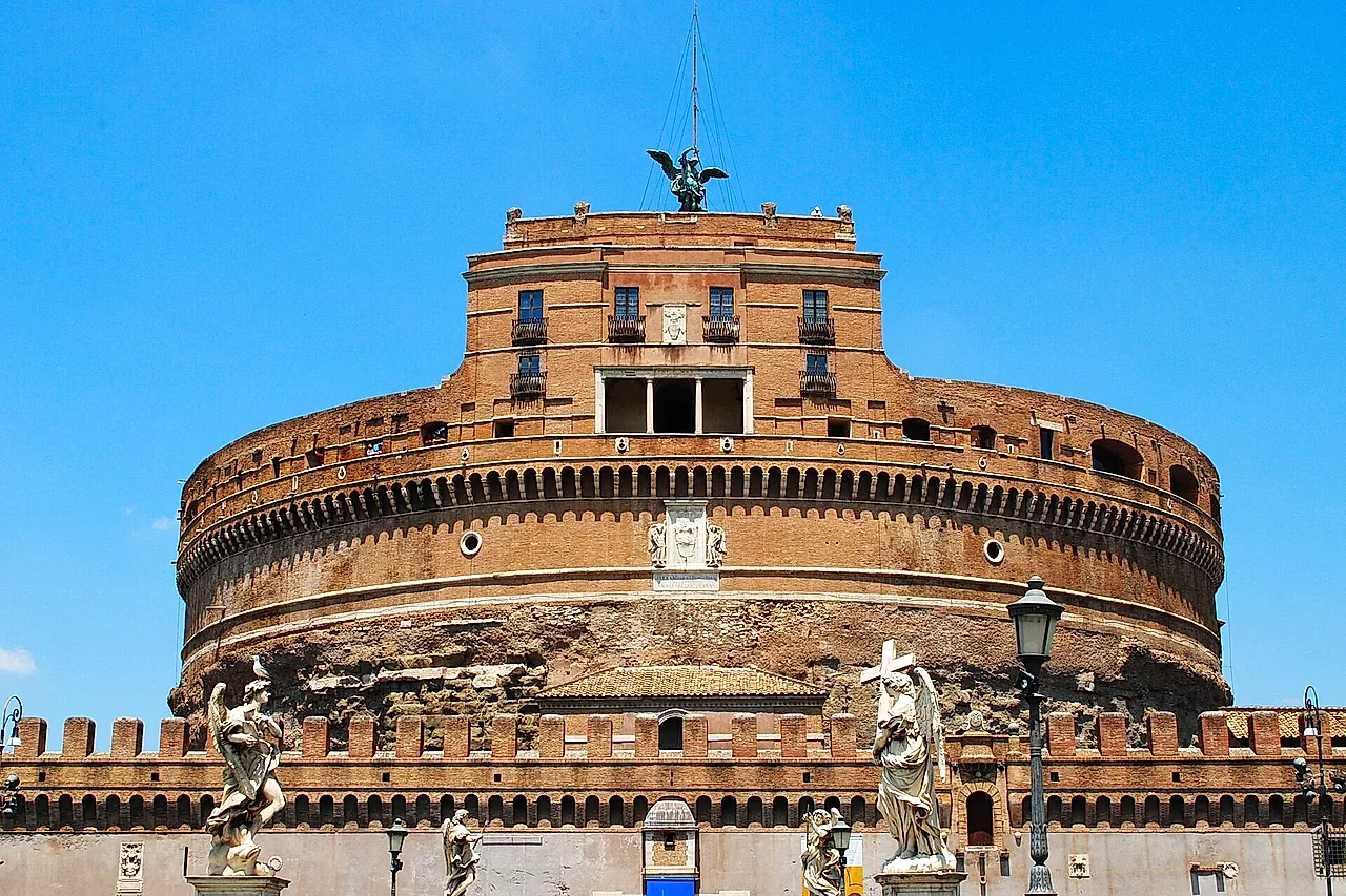 El Castillo de Sant'Angelo visto desde el Puente de Sant'Angelo sobre un cielo azul intenso: el tambor circular de ladrillo del antiguo mausoleo, el bloque superior cuadrado con el ángel de bronce en la cima y las estatuas de ángeles de mármol del puente en primer plano