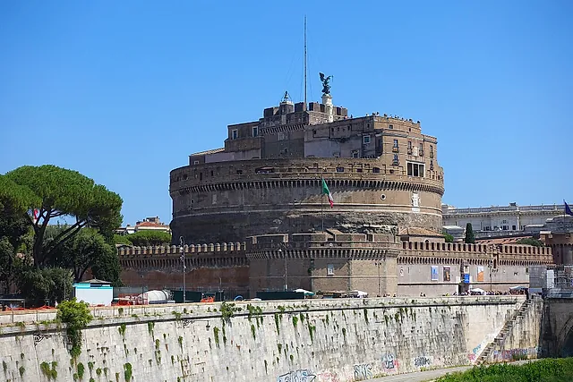 El Castillo de Sant'Angelo visto desde la ribera del Tíber en un día soleado: el macizo tambor circular y los baluartes almenados sobre el muro del río, con un pino piñonero a la izquierda