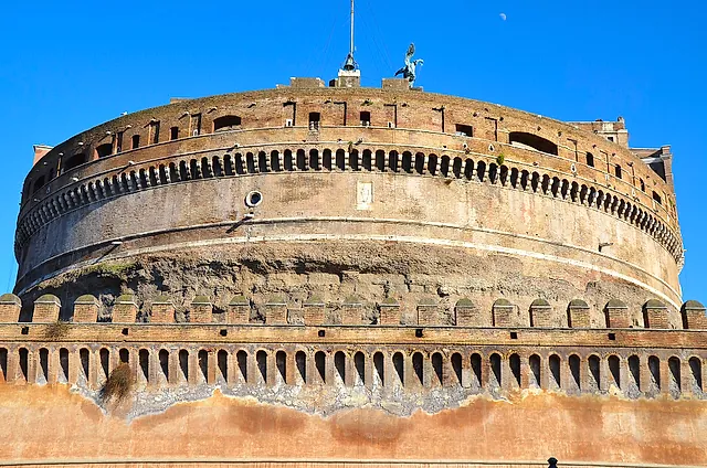 Vista cercana del enorme tambor circular del Castillo de Sant'Angelo, el antiguo mausoleo de Adriano, con sus filas de arcos pequeños y la muralla exterior almenada delante, bajo un cielo azul