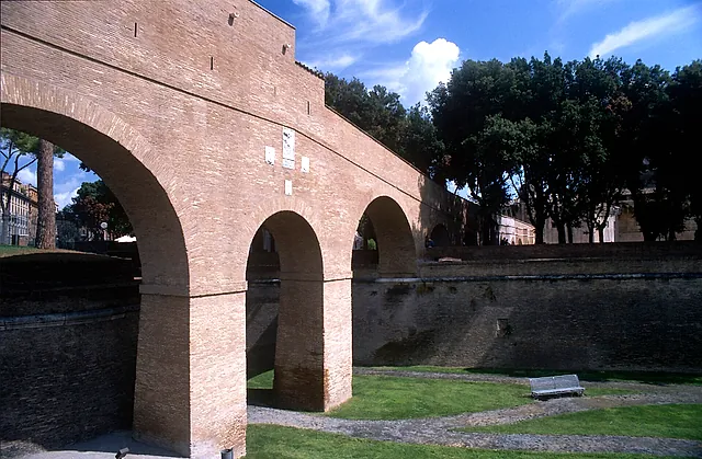 El Passetto di Borgo, el corredor elevado de ladrillo que une el castillo con el Vaticano, cruzando el foso seco cubierto de hierba sobre altos arcos entre árboles y un cielo azul