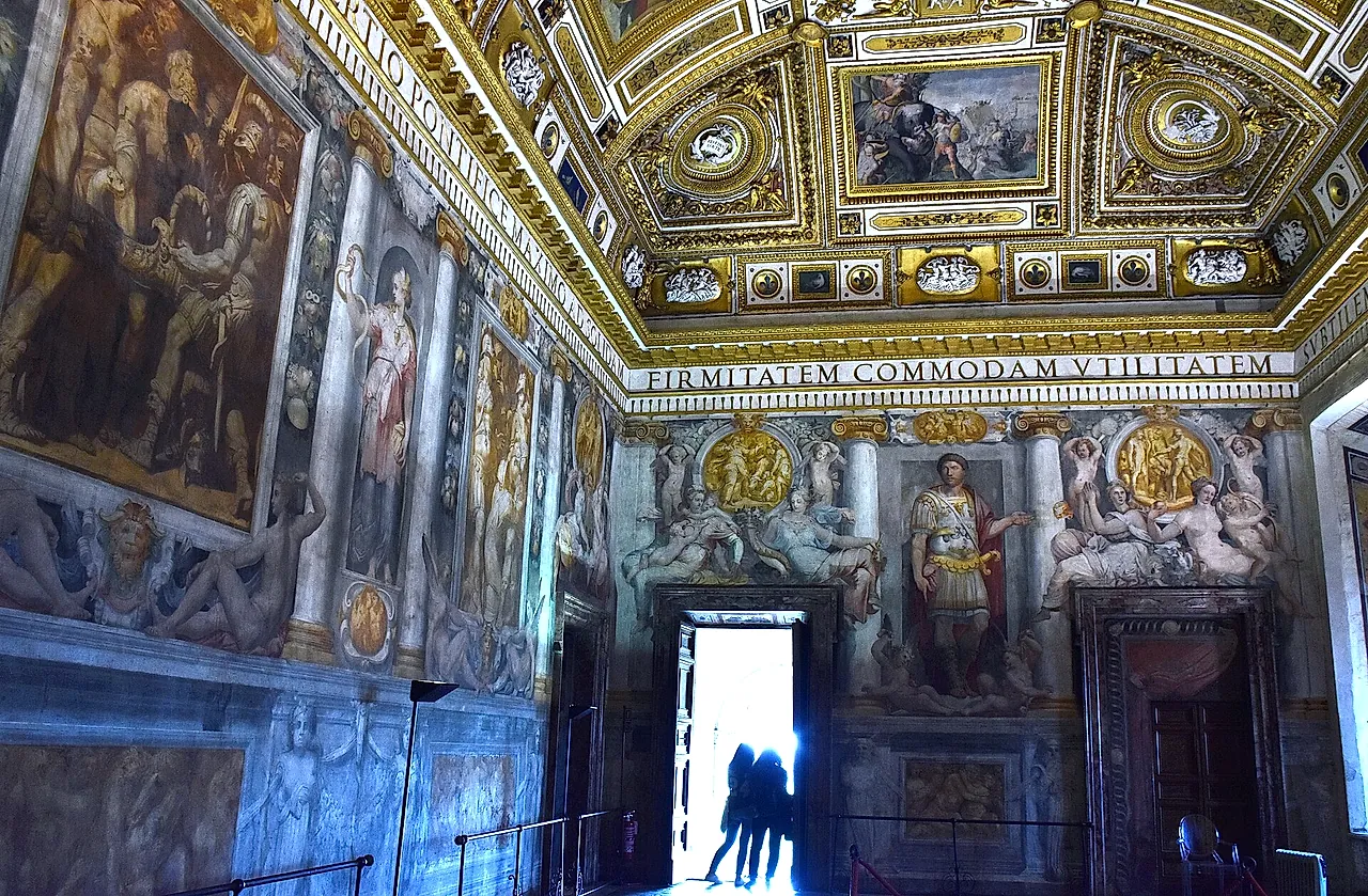 Die Sala Paolina, der Renaissancesaal der Burg, mit vergoldeter Kassettendecke, freskierten Wänden und einer hellen offenen Tür, in der zwei Besucher als Silhouetten stehen