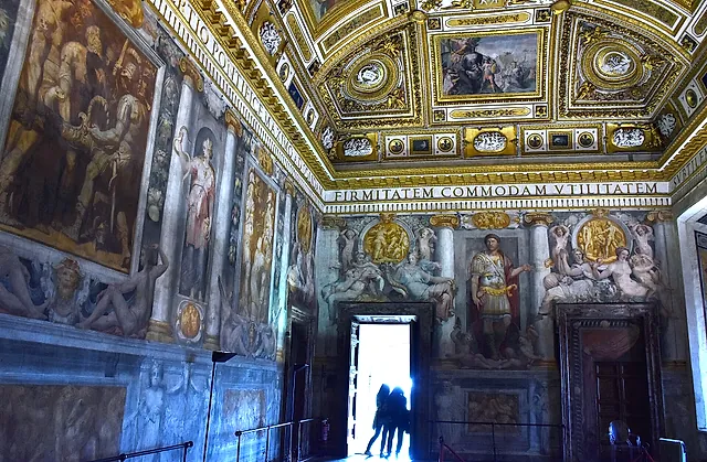 Die Sala Paolina, der Renaissancesaal der Burg, mit vergoldeter Kassettendecke, freskierten Wänden und einer hellen offenen Tür, in der zwei Besucher als Silhouetten stehen