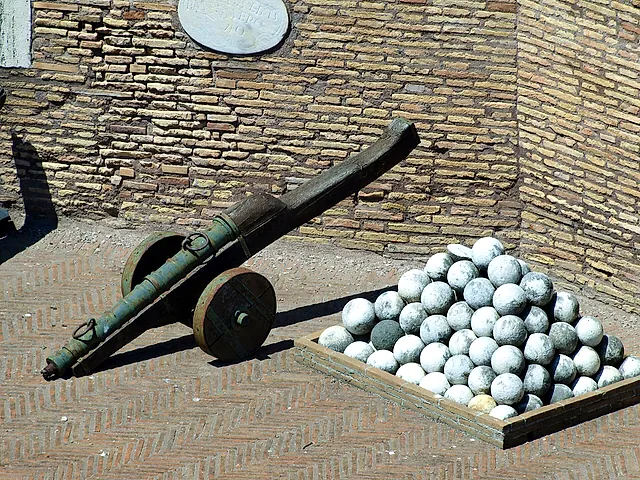 Un viejo cañón con tubo de bronce verde sobre un afuste de madera junto a un montón de balas de piedra redondas en una bandeja de madera, sobre un suelo de ladrillo en espiga delante de un muro de ladrillo