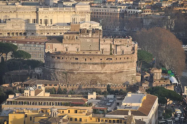Vista desde lo alto del Castillo de Sant'Angelo con luz cálida, con el tambor circular, la terraza superior donde se ven diminutos visitantes y los tejados de Roma con un gran palacio ornamentado detrás