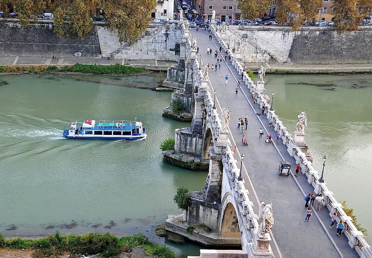 Blick von der Burg hinab auf die Engelsbrücke und den Tiber, mit einem vorbeifahrenden Ausflugsboot und Menschen, die zwischen den Engelsstatuen der Brücke gehen