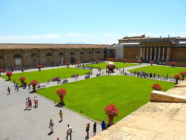 Der Cortile della Pigna in den Vatikanischen Museen von oben gesehen: grüne Rasenflächen mit roten blühenden Töpfen, Kieswege mit einigen Besuchern, die lange Galeriewand und in der Ferne eine große goldene Kugelskulptur